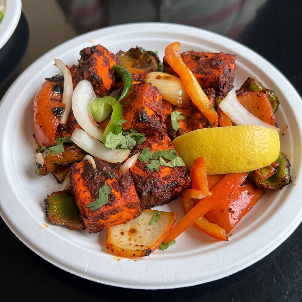 Grilled spiced paneer cubes with peppers, onions, tomatoes, cilantro, and lemon wedge on a white plate.