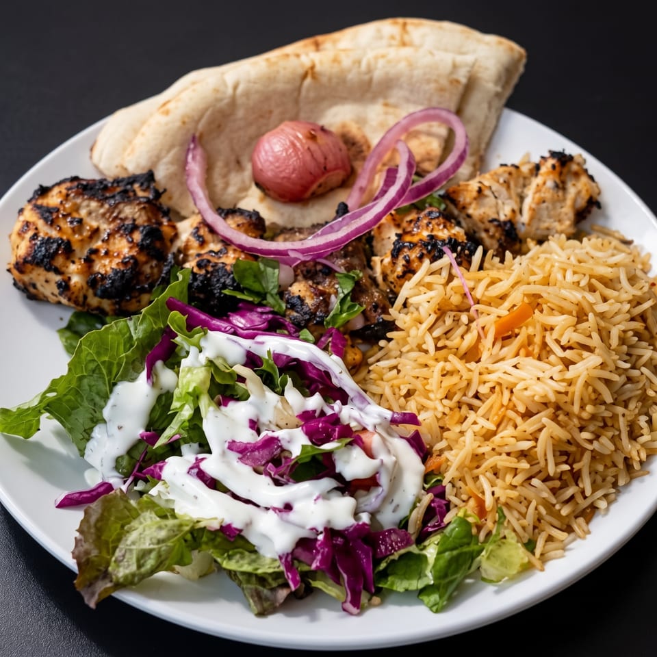 Plate of grilled chicken, seasoned rice, flatbread, red onions, salad, and creamy white sauce.