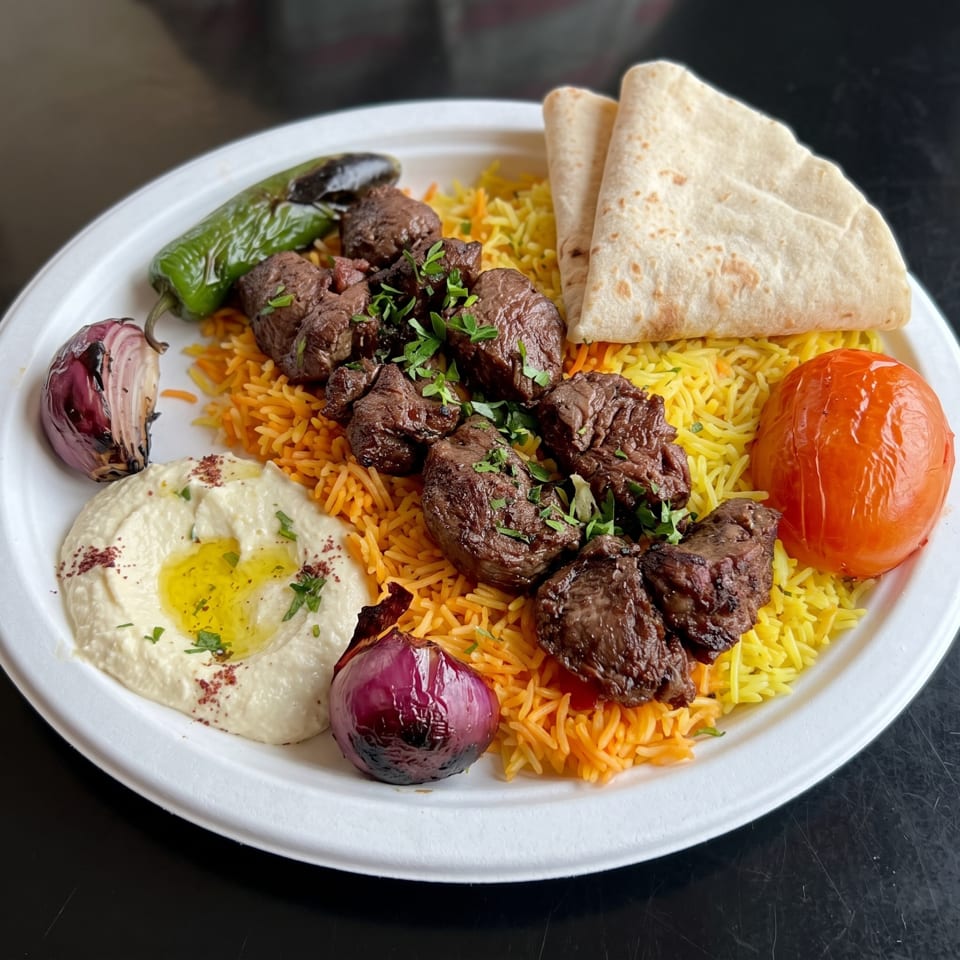 Grilled beef kebab pieces with parsley over yellow rice, pita bread, hummus, grilled vegetables, and tomato on a white plate.
